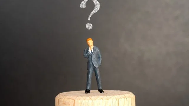 暗い灰色の背景に、スーツを着た人物の小さな像が木製の台の上に立っています。その人物は深く考え込んでいるようで、頭上には大きな疑問符が浮かんでおり、混乱や思案を象徴しています。