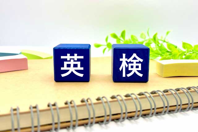 ノートの上に、日本語の文字が書かれた青いブロックが 2 つあります。左側のブロックには「英」、右側のブロックには「検」と書かれています。背景には小さな植物があります。.