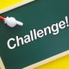 明るい黄色の背景に、白い文字で「チャレンジ！」と書かれた小さな黒板があります。赤い棒の上の白い指差し手が黒板に触れています。.