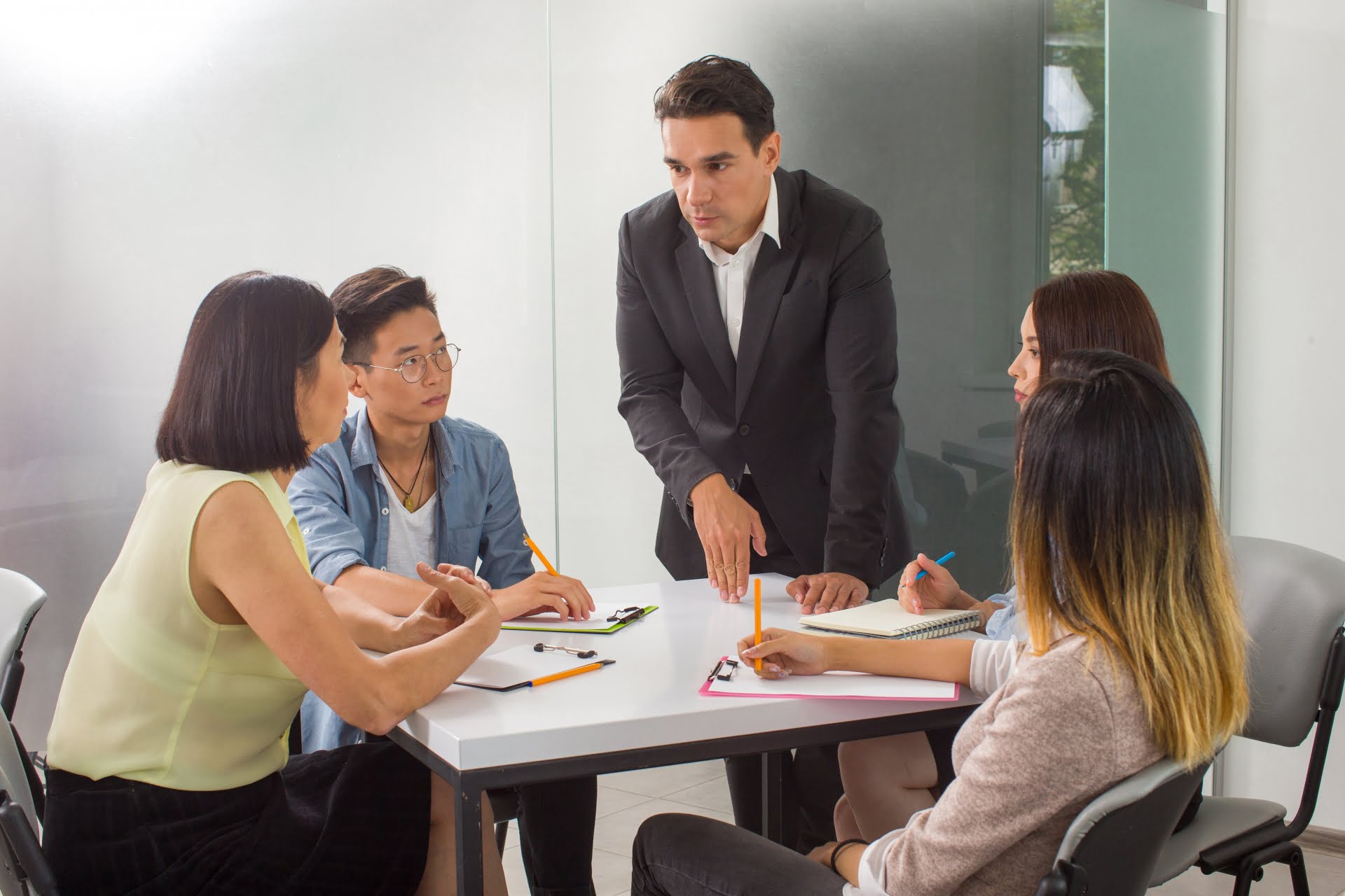 オフィスのテーブルを囲んで 5 人のグループが議論しています。1 人の男性が立って話し、他の 1 人の男性と 3 人の女性は座ってノートとペンを手に、熱心に話を聞いています。.
