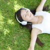 長い髪の人物が、白いノースリーブのトップスを着てヘッドフォンを着け、芝生の上に横たわっています。目を閉じており、リラックスしているか音楽を聴いている様子が伺えます。日光と影が、周囲の芝生に模様を描いています。.
