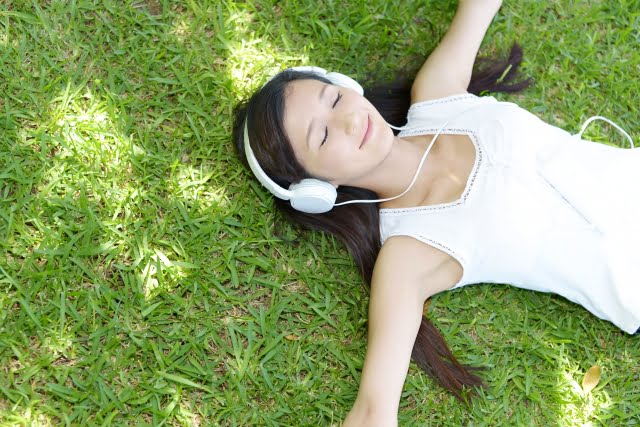長い髪の人物が、白いノースリーブのトップスを着てヘッドフォンを着け、芝生の上に横たわっています。目を閉じており、リラックスしているか音楽を聴いている様子が伺えます。日光と影が、周囲の芝生に模様を描いています。.