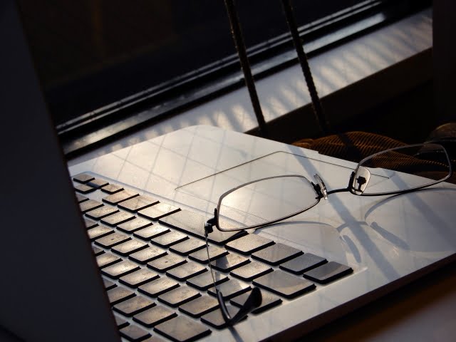 ノートパソコンのキーボードの上に眼鏡が置かれています。窓から差し込む暖かい日光がシーンを照らし、ノートパソコンとその下の表面に影と反射のパターンを作り出しています。.